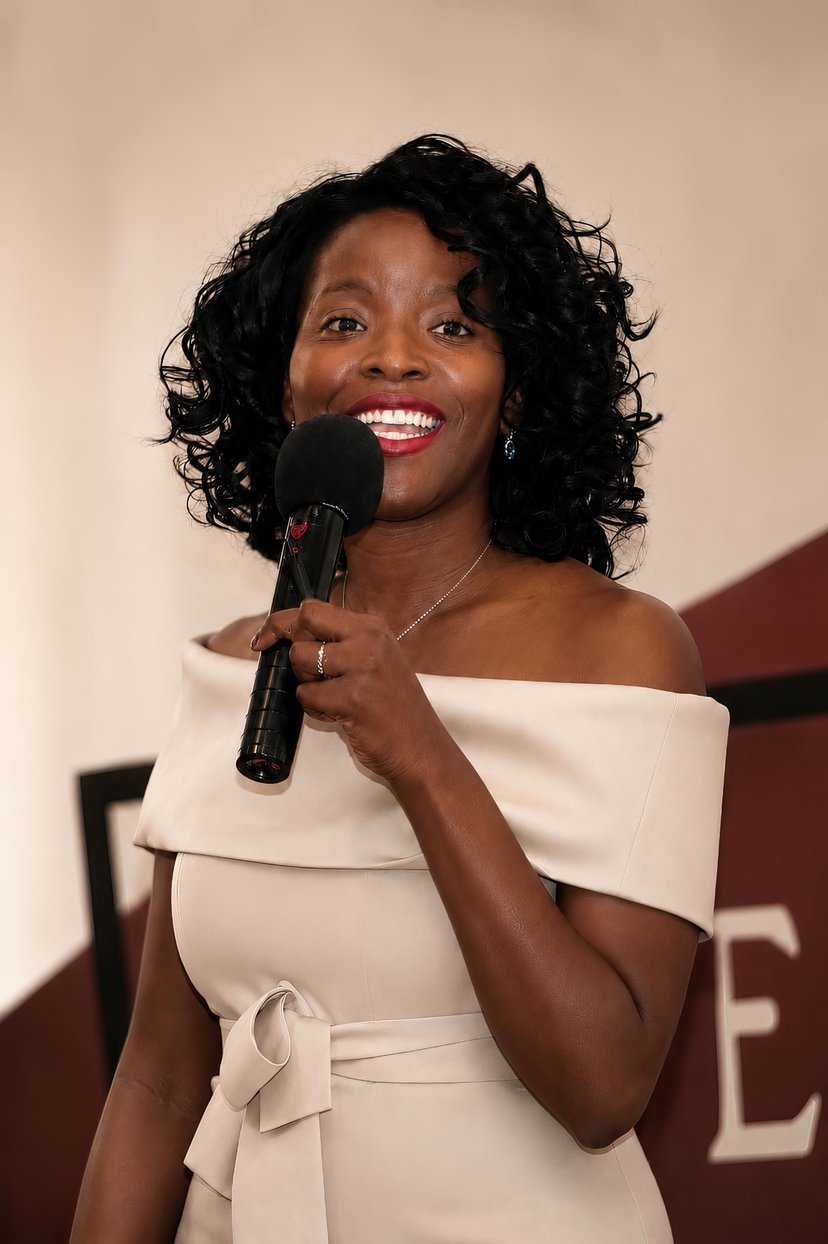 Ogo Ekweozor-Holley speaking into a microphone, smiling in a cream off-the-shoulder dress